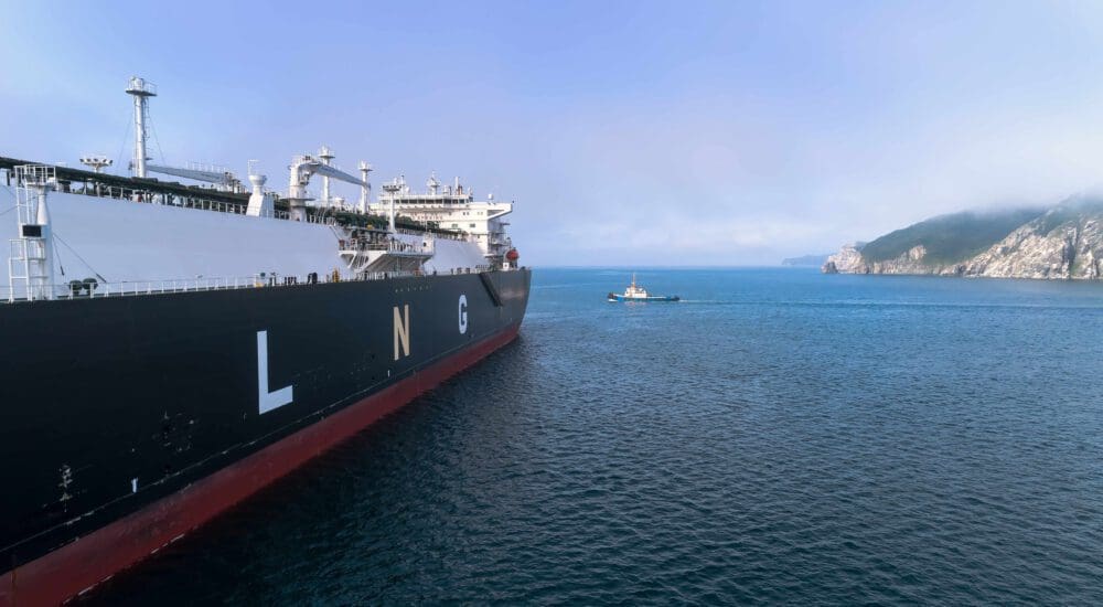 A large LNG (liquefied natural gas) carrier ship is anchored in the ocean, with a smaller boat nearby and a coastal landscape in the background.