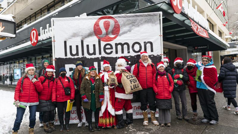 Dozens of people dressed as Santa or Mrs.Claus or elves stand by a 10 foot banner that read lululemon with its logo up top centre. The group is posing outside lululemon's store under the awning.