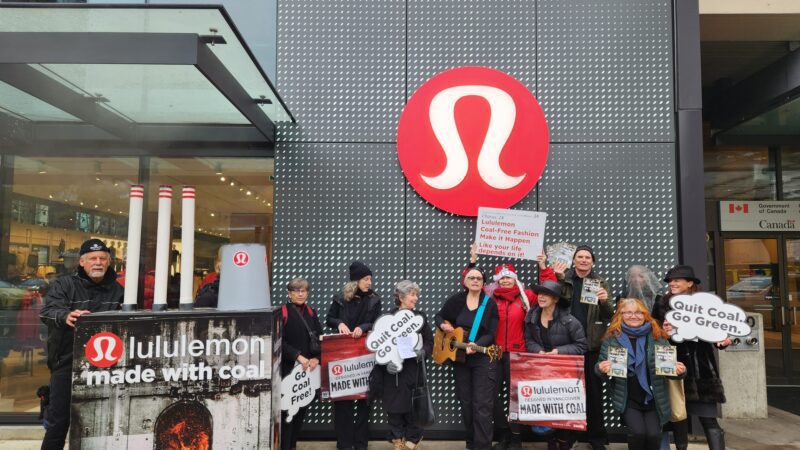 group of 11 people outside of lululemon store under a big lululemon logo carolling with posters and signs