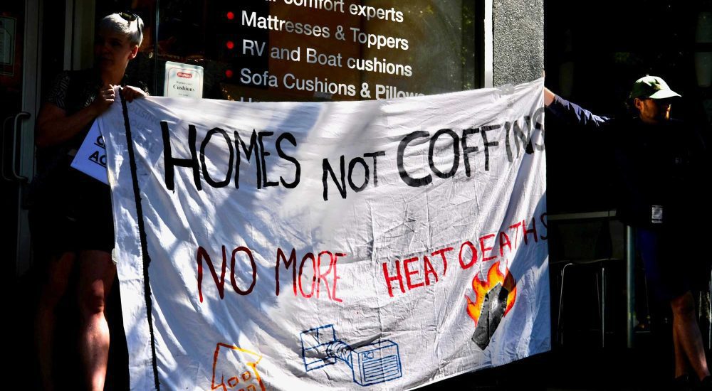 two people protesting government in action against climate crisis driven heat waves holding a banner that reads: "homes not coffins. no more heat deaths"