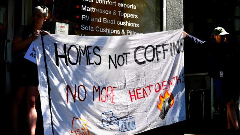 two people protesting government in action against climate crisis driven heat waves holding a banner that reads: "homes not coffins. no more heat deaths"