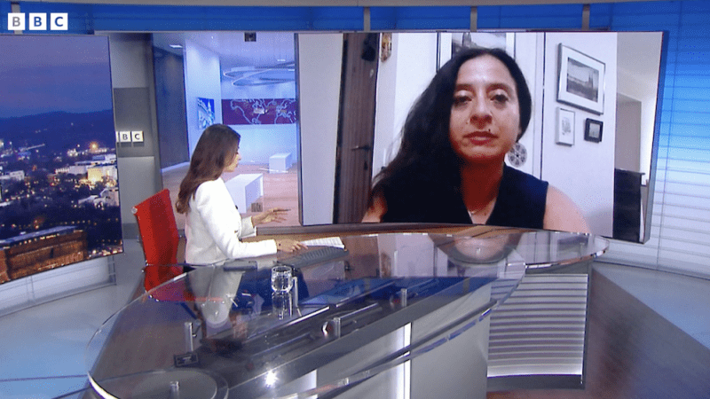 BBC newsroom with anchor seated and facing a TV screen that features Alicia Guzman