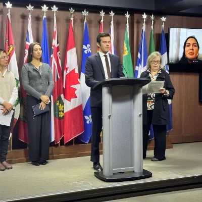 photo of four people at a press conference with one person joining remotely. one person at a podium with flag poles behind the rest of the speakers.