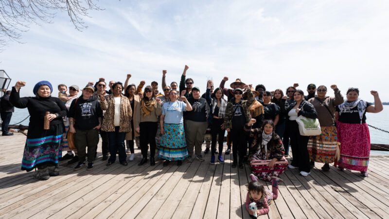 Group of 25+ Indigenous, Frontline, and Climate Delegation at the Habour Front in Toronto, many wearing traditional regalia, with fists raised.