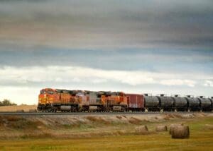 A freight train hauling tank cars filled with LNG across a rural landscape with hay bales under a cloudy sky.