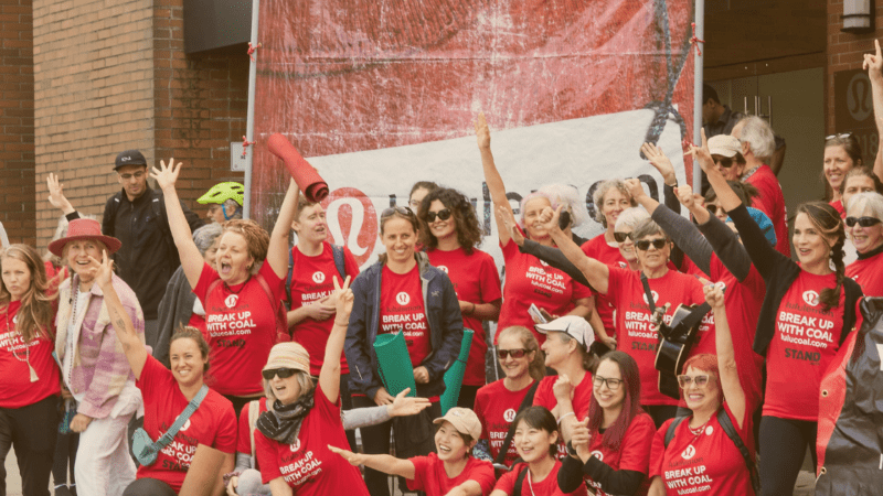A group of 20+ people in red shirts that read, "lulu, break up with coal" in white and black text hold yoga mats and pose in boisterous and happy manner in front of lululemon HQ in front of a big red banner