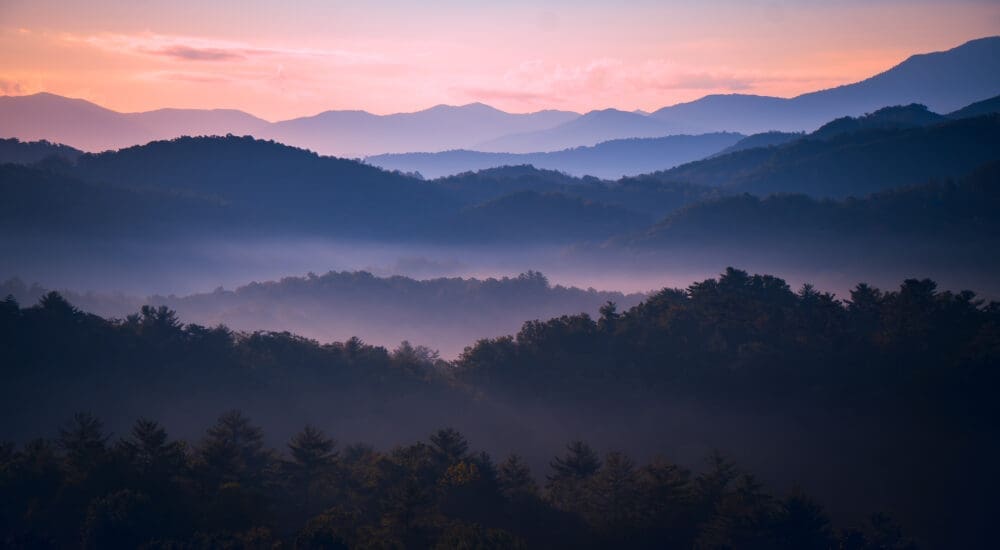 Photograph of a sunrise amongst mountains and a forest.