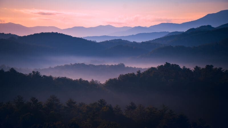 Photograph of a sunrise amongst mountains and a forest.