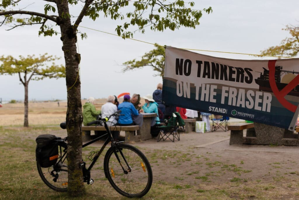 Image of sign that says No Tankers on the Fraser, a bike leaning on a tree and a picnic table full of people talking